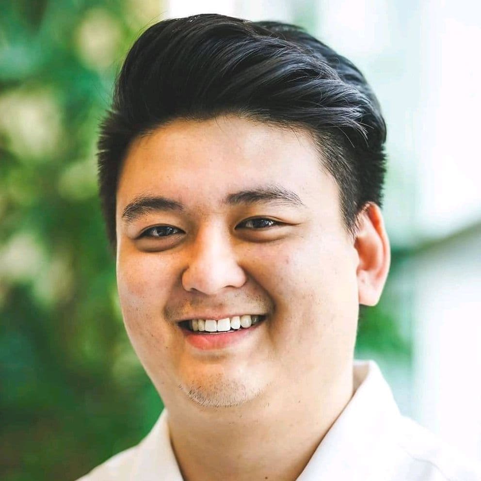 Nam Nguyen smiling, in a white shirt, with green leaves and soft daylight behind him.